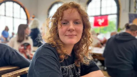 Meri has shoulder length curly red hair and a black t-shirt. She's sitting in a busy cafe with large arched windows behind her, with a Manx flag hanging in the one to the right.