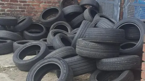 Local Democracy Reporting Service Dozens of black vehicle tyres piled beside metal alley gates between brick terraced houses.