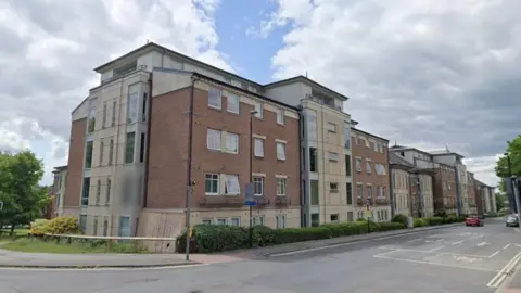 A modern red‑brick and light‑stone apartment building stretches along a street corner, with balconies, traffic lights showing red and green, a few cars on the road, and trees.