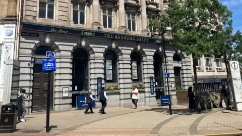 People walk by the Banker's Draft pub on a sunny day. The former bank building has grey columns and ornate detailing.