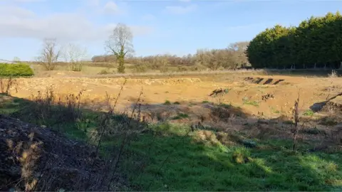 An empty field which has clearly been excavated several feet down. There are trees on the right, grass in the foreground and another field and more trees in the background.