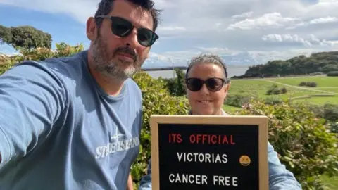 Victoria Reitze Victoria is holding a sign which reads "It's official, Victoria's cancer free". She and her husband are wearing sunglasses. In the background are The Knap gardens and Old Harbour in Barry.