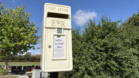 A white post box, with a panel containing black writing that reads Letters to Love Ones. The post box is in a cemetery with trees nearby, and is set against a blue sky.