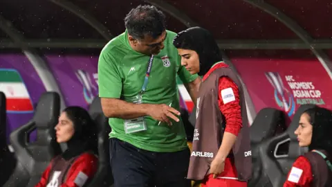 EPA Iranian coach dressed in green (on left) talks to player Mona Hamoudi, wearing red shirt with brown sleeveless top, during match as two other players sit on each side.