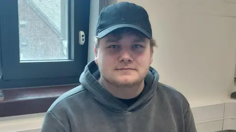 Luton Borough Council Steven Moss wearing a blue cap and a grey hoodie looking at the camera and sitting in front of a window with a wide black frame in a room with white walls.