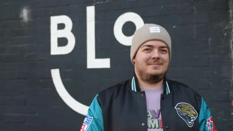 Photograph of Elliot, the owner of Bloc coffee shop in Victoria Park, along Cowbridge Road East. Elliot wars a navy and blue bomber jacket, a pink graphic t shirt and a beige beanie hat. He has blue eyes, a dark brown beard and smiles at the camera. 