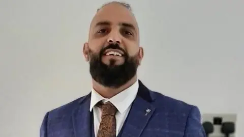 A smiling man with a black beard and a balding head. He is dressed in a navy suite with a white shirt and brown tie. 