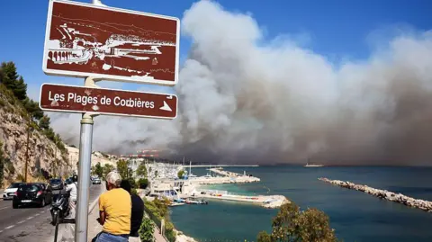 A large cloud of dense smoke fills the horizon near the Plage des Corbieres, in Marseille. In the foreground, two people are perched on a wall under the sign for the beach. 