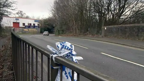A metal fence on the left side of a road has blue and white police tape knotted around it. There are trees on the right side of the road and a white building is on the left of the photo.