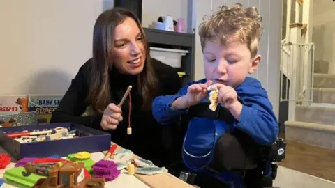A woman with long dark hair is holding a toy magnetic fishing rod and smiling at a very young boy sat next to her. He is in a wheelchair and is wearing a blue top and has blond curly hair. He is looking at a toy magnetic fish. On a table in front of them are toys. Behind them is a flight of stairs, with a toddler stairgate at the bottom.
