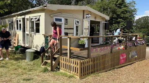 Martin Heath/BBC A wooden single-storey classroom painted in cream with white-framed windows. There are steps leading to a platform in front of the classroom. A person wearing a purple top is walking down the steps on to the grass below.