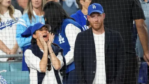 IMAGN IMAGES/Reuters Connect Harry and Meghan standing beside each in the stands of a baseball game, both wearing Dodgers hat, with Meghan cheering
