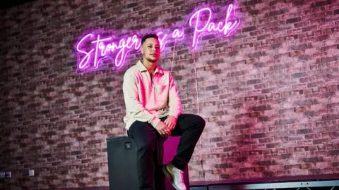 University of Wolverhampton Nate Ethan-Watson is dressed in a beige long-sleeved top and black trousers with trainers. He is sitting on a black speaker in front of a red-brick background. On the wall is a neon pink light that reads "Stronger as a pack." the pink light is reflecting on his face and clothes.