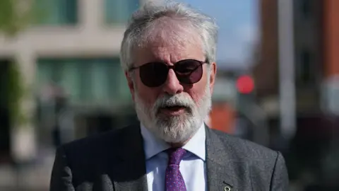 Gerry Adams with grey hair and sungalsses. He is wearing a grey suit, blue shirt and purple tie. He is outside.