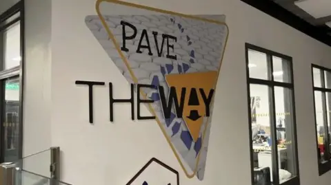 A yellow triangle is on a white wall with the words 'the way' written in black capital letters. The letter A in the word Way is stylised into an arrow logo.