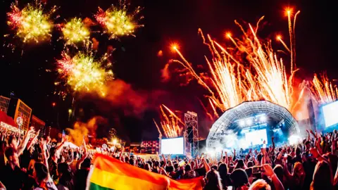 Birmingham Pride A large crowd is watching a fireworks display at a night concert. A rainbow flag is being held up in the foreground. 
