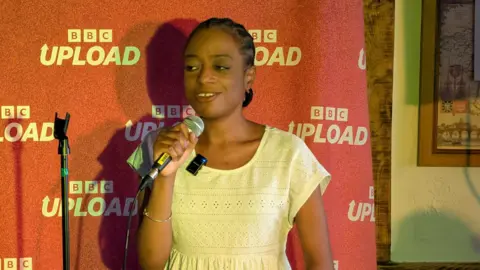 A woman talks into a microphone on stage. She stands in front of a red backdrop which says "BBC Upload" on it.