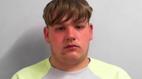 A police custody photo of a man with mid-length fair hair and dark eyes. He is wearing a yellow and grey T-shirt. He is sat in front of a grey background.