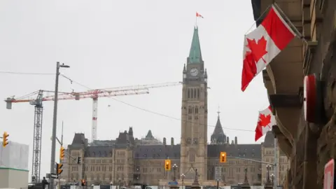 Two Canadian flags fly in the foreground with the Parliament Building in the background