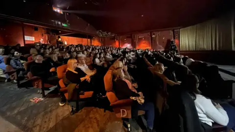 Woolton Picture House Inside Woolton Cinema with rows of people sitting on red seats watching a screen.