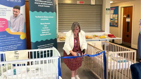 Barnsley Metropolitan Borough Council A woman in a dress and white blazer cuts a blue ribbon tied between two cots. She has short hair, styled into a bob, and wears glasses. A small buffet with different food items can be seen on the table behind her.