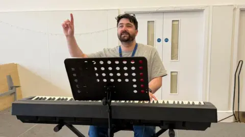 BBC/Freya Eady Brounette man with glasses on his head wearing a grey t-shirt shirt playing the keyboard and pointing up on stage.