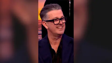 David McCormack, the voice of Bandit Heeler, smiles. He wears black glasses and has swept back grey hair.