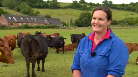 A farmer - a woman in her 40s wearing a blue shirt with rolled up sleeves - stands in a field surrounded by calves on a gloomy day. A farmhouse can be seen in the distance.