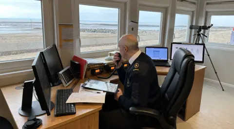 NCI Minehead A man wearing a navy blue watch-keeper uniform using a radio to communicate with someone. He is looking out of the window out to sea.
