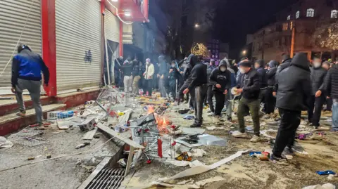 A group of people in face masks and hoods gather around burning debris outside shuttered shops in Kermanshah on Thursday.