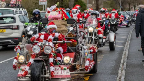 Del Hickey A group of people all dressed as Santa drive in procession on motorbikes. Many of the bikes, especially the one at the front, are covered in tinsel and other festive decorations. 