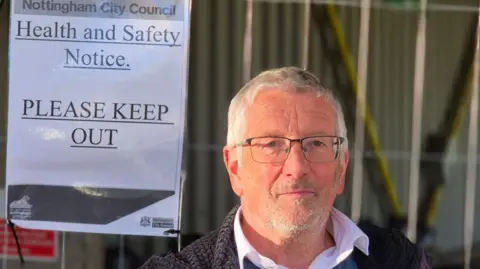 BBC Notts AC Chairman, Mick Robinson, wearing a white shirt under a blue v-neck jumper and navy coat. He is standing in front of a wire fence with a health and safety notice on it.