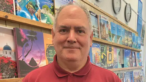 Adele Forrest/BBC A man with short grey hair wearing a red polo shirt stands in front of a wall or travel brochures