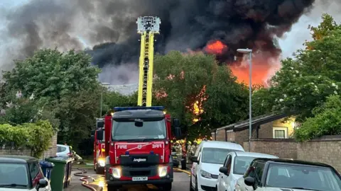 Cambridgeshire Fire and Rescue A specialist fire vehicle which has a yellow ladder extending from it. There are houses on either side and trees behind. Beyond that can be seen black smoke and orange and yellow fire