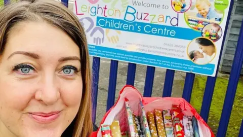 Holly King-Mand Holly King-Mand has dark blonde shoulder-length hair and pale blue eyes and is smiling at the camera. She is carrying a bag of books, individually wrapped with Christmas wrapping paper. She is outside Leighton Buzzard children's centre.