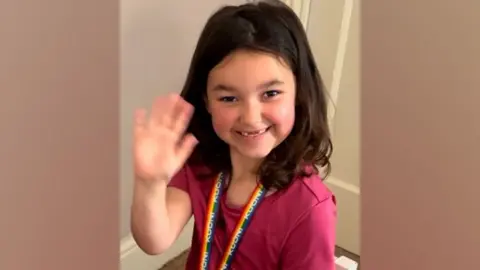 Bebe's Hive Bebe is pictured sitting in a little white wooden chair. She is smiling and waving with her right hand. She has long brown hair and is wearing a pink top and pink patterned leggings.