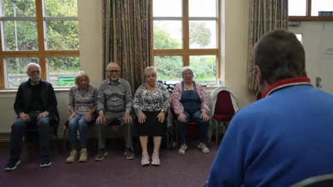 Five people sit in chairs against a wall with two windows in it. They are all sitting upright with their hands on their knees. They are being led in breathing exercises by Jim who has his back to camera in the corner of the frame. 