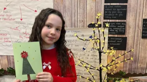 The image shows Seren standing indoors, holding a handmade Christmas card. The card is green with a decorated Christmas tree design in the center, topped with a red bow. The child is wearing a bright red sweater with two white ribbon bows on the front. Behind them, the background includes educational posters with handwritten notes and diagrams, a decorative tree with yellow lights or flowers, and a wooden-themed wall display. There are also books and classroom materials visible, suggesting this is a festive activity in a school setting.
