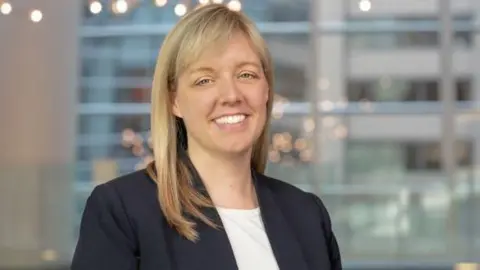 A woman with blonde hair smiling at the camera. She is wearing a navy jacket and white top.