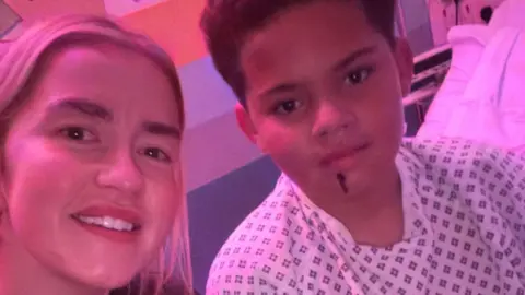 A woman and a boy in a selfie under red light in a hospital room with brightly coloured walls. The boy is wearing a white and blue hospital gown and sitting alongside his mum. A large gash can be seen on his chin. He has brown hair. The woman is blonde, wearing a black Nike sweatshirt and smiling.