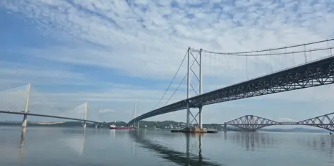 John Gallagher The Queensferry Crossing, Forth Road Bridge and Forth Rail Bridge spanning across the water. The reflections of the bridges are in the water and there are clouds in the sky.