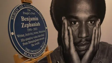 A plaque rests on a wooden frame with a black and white photo of Zephaniah as a younger man with his hands on his face.
