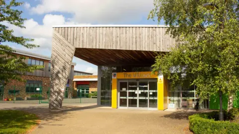 The entrance to a modern school building. Columns are painted yellow around a glass door, with a large wood clad pillar and roof. Willows is written in silver letters above the door, with reception written on the glass. Other school buildings can be seen to the left, with trees and bushes lining the path.