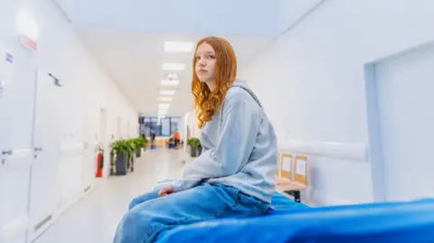 Young person with long red hair, wearing a grey hoodie and jeans, sits on a blue hospital trolley in a white corridor behind her and bright lights above. 