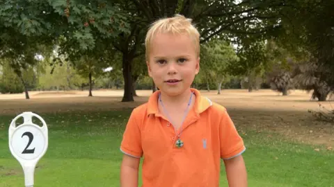 BBC/Lilah Parsons Jude is wearing an orange polo shirt stands on a putting green next to a white marker displaying the number “2”, with mature trees and open parkland visible behind.