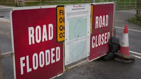 A close up of a red sign saying road flooded road closed in white letters. It blocks one side of the road. 