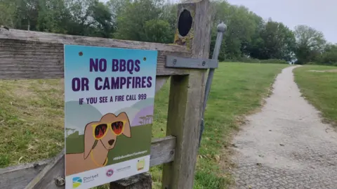 Litter Free Dorset An open wooden gate on a footpath. Attached to the gate is a sign which says no BBQs or camp fires, if you see a fire dial 999. It also bears a cartoon of a brown dog which is wearing yellow sunglasses with flames reflected in the lenses.