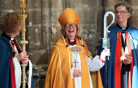 Stefan Rousseau / PA Media Dame Sarah wears a robe and holds a staff while laughing and smiling outside the cathedral after the ceremony.