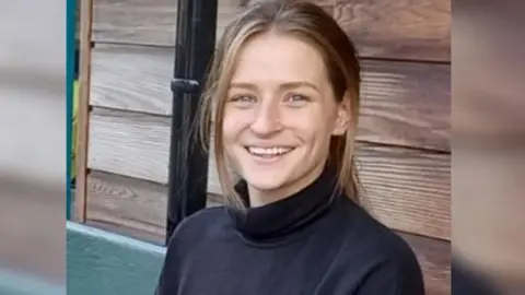 Family handout A young woman with long hair in a pony tail and wearing a black turtle neck smiles at the camera. She is sitting in front of a shed.