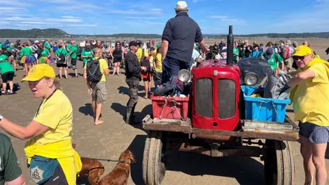 A busy beach scene is shown on a bright, sunny day with a clear blue sky streaked by thin white clouds. The photograph is taken at ground level on wet, firm sand, and the view centres on an old red tractor surrounded by a large crowd of people.